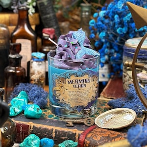Op de afbeelding: Een glazen kaars met blauwe was en een paarse, slagroomachtige topping, versierd met een blauwe zeemeerminstaart. De kaars is voorzien van het opschrift "Mermaid Tears" in een vintage stijl. De kaars staat tussen andere decoratieve items.