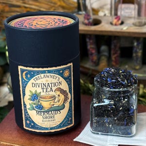 May include: A dark blue cylindrical container of Trelawney's Divination Tea, with a label featuring a mermaid and blueberries. A glass jar of loose leaf tea sits beside it. The tea is labelled "Mermaid's Shore Blueberry".