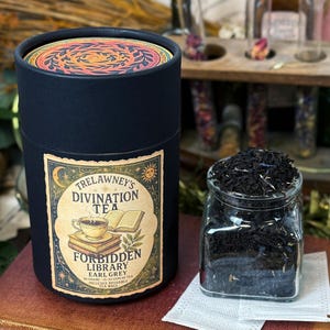 May include: A black cylindrical container of Trelawney's Divination Tea, with a label featuring a teacup and books. Beside it, a glass jar holds loose-leaf tea. The container's lid has a colorful, circular design. The tea is labeled "Forbidden Library Earl Grey".