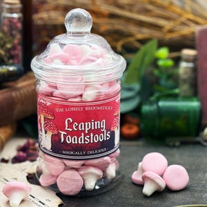 Peut inclure: Un bocal en verre transparent rempli de bonbons roses et blancs en forme de champignon. Le bocal porte une étiquette rouge avec le texte "Leaping Toadstools" et "Magically Delicious". Les bonbons sont éparpillés.