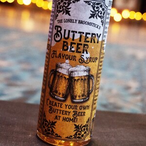 May include: A brown glass bottle with a yellow label that says "The Lonely Broomstick's Buttery Beer Flavor Syrup". The label has a vintage design with illustrations of two beer mugs and the text "Create your own Buttery Beer at home!"