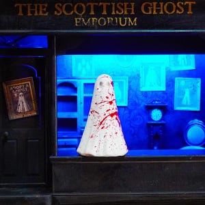 May include: A model shopfront with the words "THE SCOTTISH GHOST EMPORIUM" above the window. A white ghost figure with red splatters is in the window. Inside, framed pictures and a clock are visible. A sign that says "ADOPT A GHOST" hangs on the door.