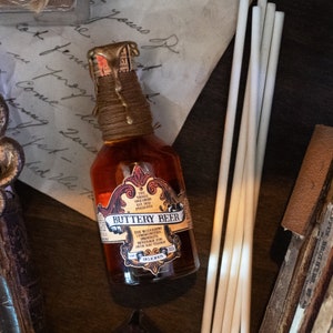 May include: A glass bottle of Buttery Beer with a gold and brown label. The label features a stylized unicorn and the text "The Triple Unicorns Est. 1701 Presents Buttery Beer". The bottle is sitting on a piece of paper with writing on it.