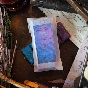 Op de afbeelding: Een wax melt in een doorzichtige plastic zak met een label waarop staat "The Lonely Broomstick Wax Melt Collection Mermaid Tears". Het label heeft een paars-blauwe ombre-effect. De tekst op het label luidt: "Ontdek "Mermaid Tears", waar de verfrissende geur van watermeloen een vleugje oceanische schoonheid aan je ruimte toevoegt. Dompel jezelf onder in de zoetheid van rijpe watermeloen, die visioenen oproept van zoute golven en fantasierijke zeemeerminverhalen."