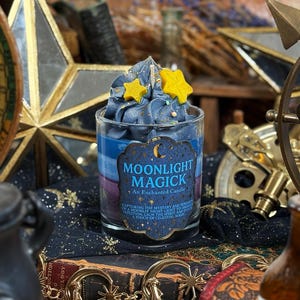 Puede incluir: Una vela de cristal "Moonlight Magick" con un diseño azul y morado en capas, rematada con cera azul y detalles de estrellas amarillas. La vela se encuentra entre una decoración de temática celestial, incluido un adorno en forma de estrella.