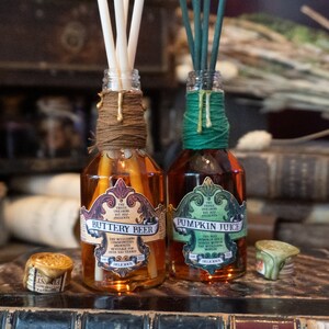 May include: Two glass bottles with brown and green twine wrapped around the necks. The bottles contain a brown and amber liquid and have labels that read "Buttery Beer" and "Pumpkin Juice". The labels are decorated with ornate designs and text that reads "The Three Unicorns Ent. Presents."