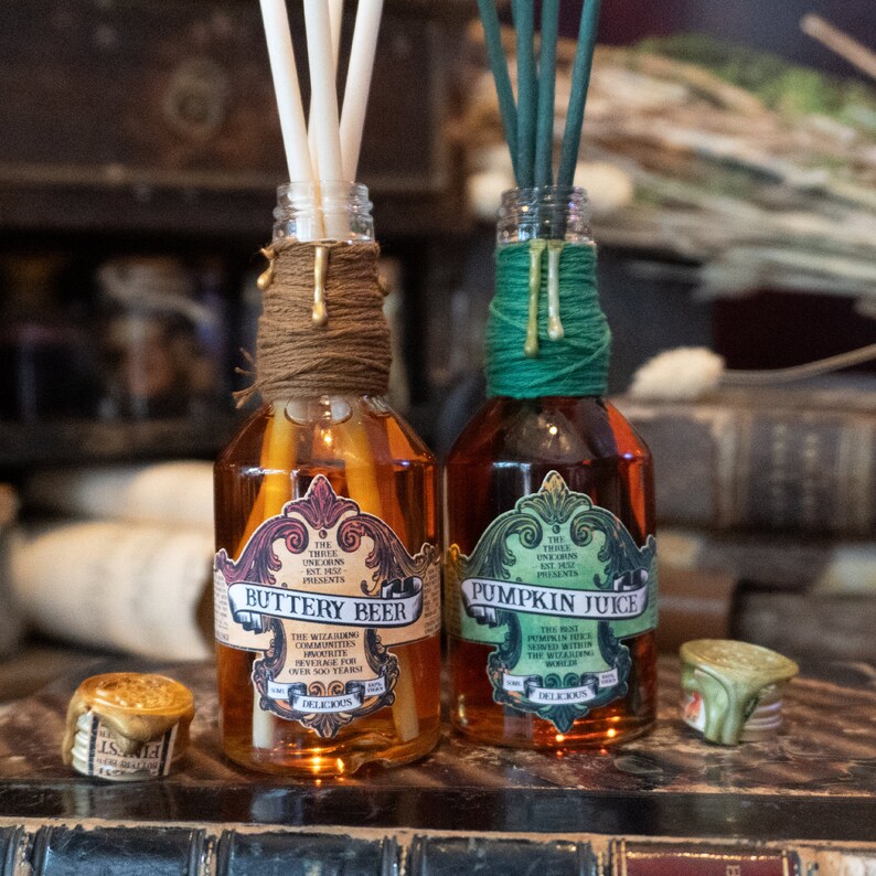 May include: Two glass bottles with brown and green twine wrapped around the necks. The bottles contain a brown and amber liquid and have labels that read "Buttery Beer" and "Pumpkin Juice". The labels are decorated with ornate designs and text that reads "The Three Unicorns Ent. Presents."