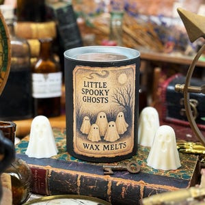 May include: A black candle holder with a label that reads "Little Spooky Ghosts Wax Melts" featuring a spooky illustration of ghosts. Several white ghost-shaped wax melts are arranged around the candle. The scene is set on a vintage book.
