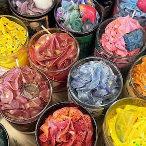 May include: A collection of decorative candles in glass jars, each topped with colorful, textured wax resembling frosting. The candles are in various colors, including pink, blue, yellow, and red, with glitter accents. Some candles have decorative elements.