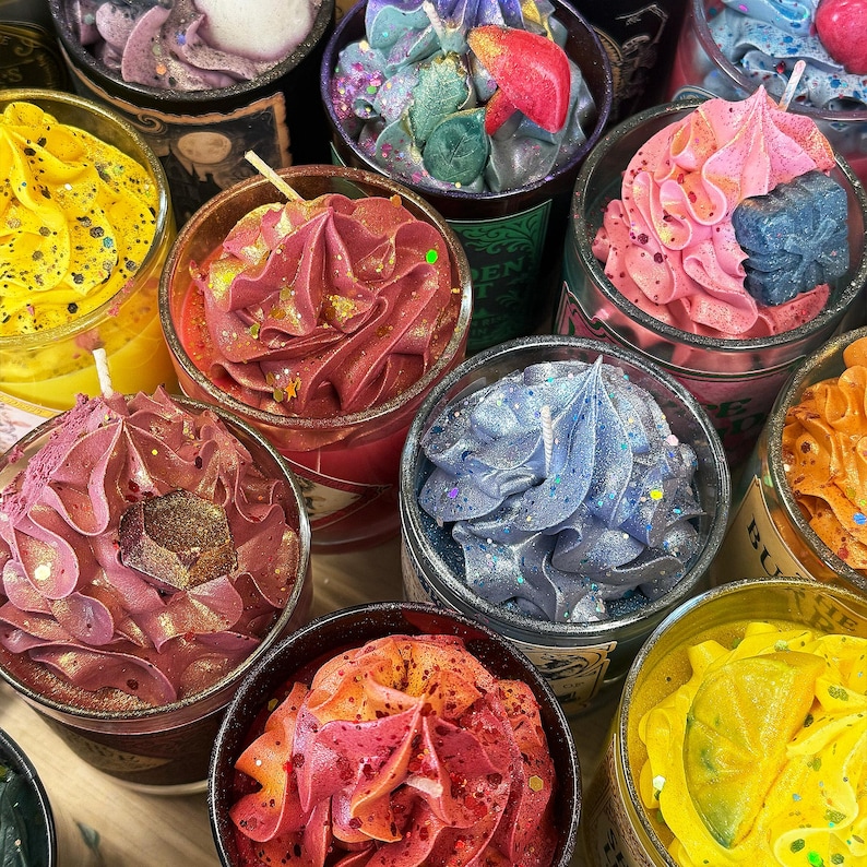 May include: A collection of decorative candles in glass jars, each topped with colorful, textured wax resembling frosting. The candles are in various colors, including pink, blue, yellow, and red, with glitter accents. Some candles have decorative elements.