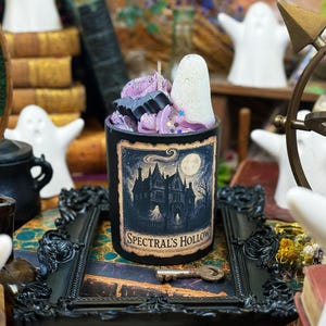 Peut inclure: Une bougie noire avec une illustration effrayante et le texte "Spectral's Hollow". La bougie est surmontée de cire violette, d'un fantôme blanc et d'une chauve-souris noire. Elle repose sur un plateau décoratif noir avec une clé.