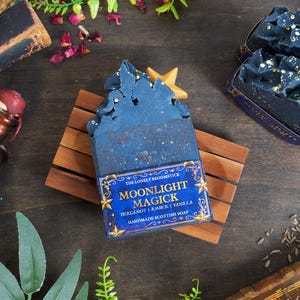 Puede incluir: Un jabón escocés artesanal con una etiqueta azul y dorada que dice "Moonlight Magick". El jabón es azul oscuro con un acento en forma de estrella. El jabón está sobre un jabonera de madera. Otros artículos incluyen una botella vintage y libros antiguos.