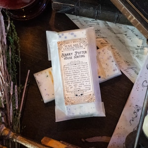 Könnte beinhalten: Ein weißes Wachs-Schmelzstück mit blauem Glitzer, verpackt in einer durchsichtigen Plastiktüte. Das Wachs-Schmelzstück ist mit "The Lonely Broomstick Wax Melt Collection Harry Potter House Sorting" beschriftet. Das Etikett hat ein schwarzes und goldenes Design mit einem Hogwarts-Hauswappen.