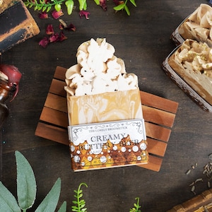 May include: A bar of handmade soap with a creamy, layered appearance, topped with a decorative, light-colored design. The soap rests on a wooden soap dish, with the label reading "Creamy Butter Beer". Additional soaps and vintage items are in the background.