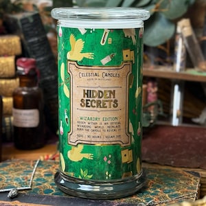 Puede incluir: Un tarro de vela de vidrio verde con tapa transparente. La etiqueta dice "Celestial Candles Hidden Secrets Wizardry Edition". El tarro está decorado con pájaros amarillos y elementos mágicos. La vela es de 600g y dura 90 horas.