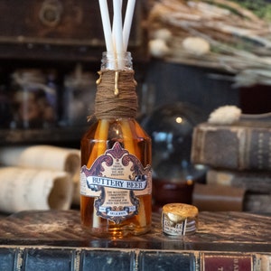 Puede incluir: Una botella de vidrio con una etiqueta marrón que dice "Buttery Beer" y presenta un diseño dorado. La botella está llena de un líquido dorado y tiene palitos difusores blancos que sobresalen de la parte superior.