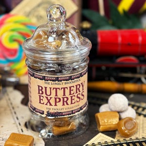 May include: A glass jar filled with sweets, labelled "BUTTERY EXPRESS", with a toy train in the background. The jar has a lid and is surrounded by other sweets and a colourful lollipop. The label also says "The Lonely Broomstick" and "The Trolley Collection".