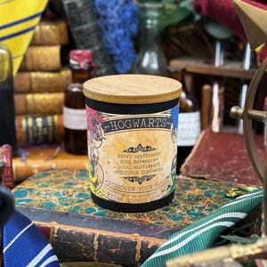 Può includere: Un barattolo di candela nero con coperchio in legno, etichettato "HOGWARTS". L'etichetta presenta lo stemma di Hogwarts e i colori delle case. Il barattolo è appoggiato su una pila di libri antichi, con altri oggetti vintage sullo sfondo.