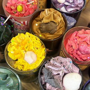 May include: A collection of decorative candles in various colors, including pink, yellow, and purple. Each candle is in a black container and topped with unique designs, such as a frog, crystal, and bat shapes. Some candles have glitter accents.