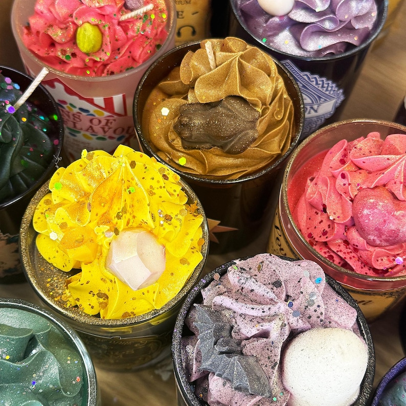 May include: A collection of decorative candles in various colors, including pink, yellow, and purple. Each candle is in a black container and topped with unique designs, such as a frog, crystal, and bat shapes. Some candles have glitter accents.