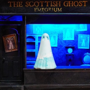 Op de afbeelding: Een miniatuur etalage met de tekst "THE SCOTTISH GHOST EMPORIUM". Een witte spookfiguur met blauwe accenten staat in de etalage. Een ingelijst bord leest "ADOPT A GHOST". Het interieur is blauw verlicht.