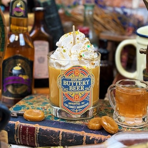 May include: A candle in a clear glass with a label that reads "Buttery Beer" and topped with whipped cream and sprinkles. The candle sits on a vintage book with caramel candies nearby. Other bottles and a mug are in the background.