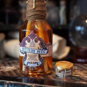 May include: A glass bottle of Buttery Beer with a gold-colored label and a brown twine tied around the neck. The label features an ornate design and the text "The Three Unicorns Est. 1452 Presents Buttery Beer The Wizarding Communities Favourite Beverage for Over 500 Years! Delicious". A small gold-colored bottle stopper is also visible.