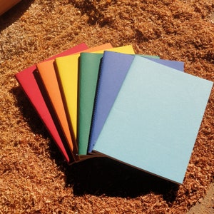 May include: A set of seven notebooks in rainbow colours, stacked on top of each other. The notebooks are all the same size and have a simple design.