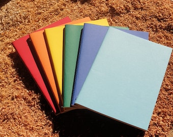 May include: A set of seven notebooks in rainbow colors, stacked on top of each other. The notebooks are all the same size and have a simple design.