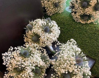 May include: Five white baby's breath bouquets with blue thistle accents. The bouquets are arranged on a dark brown surface and a green surface.