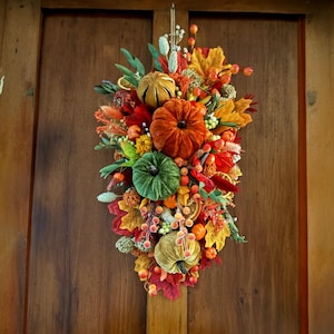 Könnte beinhalten: Ein herbstliches Türschild mit Kürbissen in Orange, Grün und Gelb. Die Anordnung umfasst bunte Blätter, Beeren und andere dekorative Elemente und schafft eine festliche Herbstdekoration.