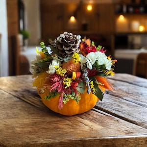 May include: A decorative orange pumpkin centrepiece featuring a vibrant arrangement of dried flowers, including white carnations, yellow statice, red accents, green foliage, and a large brown pinecone centred at the top of the floral display.