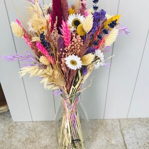 Pu&ograve; includere: Un mazzo di fiori secchi in una bottiglia di latte di vetro trasparente. Il mazzo comprende lavanda secca, grano e margherite in tonalit&agrave; di rosa, viola e crema.