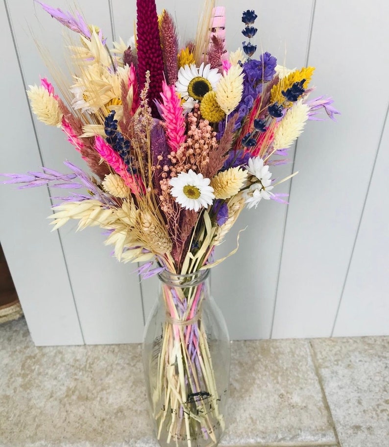 Pu&ograve; includere: Un mazzo di fiori secchi in una bottiglia di latte di vetro trasparente. Il mazzo comprende lavanda secca, grano e margherite in tonalit&agrave; di rosa, viola e crema.