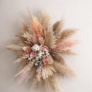 May include: A large, circular floral arrangement featuring dried pampas grass, blush-colored roses, and various other dried flowers. The arrangement includes a mix of textures and colors, with a neutral and natural aesthetic.