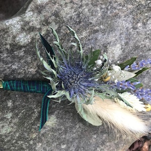 Puede incluir: Un pequeño ramo de flores secas con un cardo azul, lavanda y hierba de la pampa blanca, atado con una cinta verde y negra.