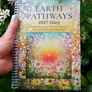 May include: A spiral-bound 2027 diary titled Earth Pathways. The cover features a vibrant, colorful illustration of a sunlit meadow with flowers and sparkles. The text reads: Earth Pathways 2027 Diary, Celebrating our interconnection with the Earth and each other.