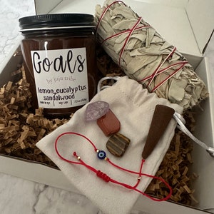 May include: A white box filled with brown packing paper contains a brown candle with the label "Goals by Juju Tribe Lemon Eucalyptus Sandalwood 7.3 oz (200g)", a white sage smudge stick tied with red string, a small white pouch with a brown incense cone, a red string bracelet with a blue evil eye charm, and four small crystals: amethyst, carnelian, tiger's eye, and red jasper.