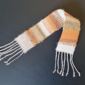 Op de afbeelding: Een geweven sjaal met een wit, bruin en oranje gestreept patroon. De sjaal heeft franjes aan beide uiteinden.
