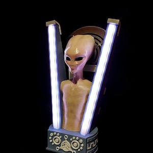 May include: A grey alien bust with large eyes and a smooth, pale skin tone. The bust is illuminated by two bright white lights on either side. The bust is on a base with a circular design.