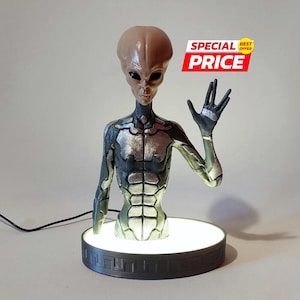 May include: A detailed sculpture of an alien figure with a gray-brown head and dark eyes, wearing a black and silver suit. The figure is mounted on a round, illuminated base. The text "SPECIAL PRICE BEST OFFER" is visible.
