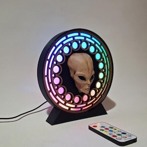 May include: A black and brown alien-themed LED lamp with a remote. The lamp features a detailed alien head in the center, surrounded by a circular frame with colorful LED lights. The lamp is on a black stand.