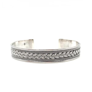 Könnte beinhalten: Ein silbernes Armreif-Armband mit geflochtenem Design. Das Armband hat ein breites Band mit einem detaillierten Muster aus ineinander verwobenen Strängen. Das offene Design ermöglicht ein einfaches Tragen. Das Armband besteht aus glänzendem Metall.