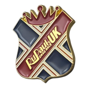 May include: A shield-shaped pin with a gold border, featuring a crown at the top. The pin has a red and navy blue color scheme with the text "RuPaul's Drag Race UK" in gold. The design incorporates geometric shapes.