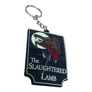 Op de afbeelding: Een zwarte rubberen sleutelhanger in de vorm van een bord met de tekst "The Slaughtered Lamb" in wit. Het bord toont een cartoonafbeelding van een wolfskop met een bloederige bek en een maan op de achtergrond.