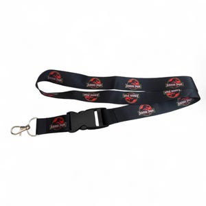 May include: A black fabric lanyard featuring a repeating Jurassic Park logo pattern in red, white, and black. The lanyard includes a black plastic side-release buckle and a silver-toned metal swivel lobster claw clasp for attaching items.