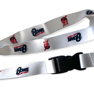 Puede incluir: Lanyard blanco con un clip de plástico negro y un gancho de metal plateado. El lanyard tiene un patrón repetido de la cara de David Bowie y la palabra "BOWIE" en letras azules y rojas.