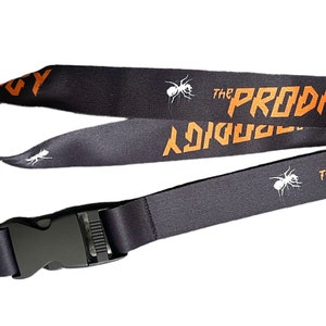 Puede incluir: Lanyard negro con un clip plateado y una hebilla de plástico negro. El lanyard tiene el texto "The Prodigy" impreso en naranja, junto con un gráfico de una hormiga blanca.