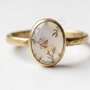 May include: A gold ring featuring an oval-shaped dendritic agate gemstone with dark, fern-like inclusions set in a smooth bezel. The band has a textured, hammered finish, and the word gems is partially visible on the surface of the stone.