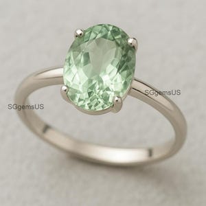 May include: A silver ring with a large, oval-shaped, pale green gemstone. The gemstone is held in place by four prongs, and the band is polished. The ring is shown against a plain backdrop, emphasizing the gemstone's colour and clarity.